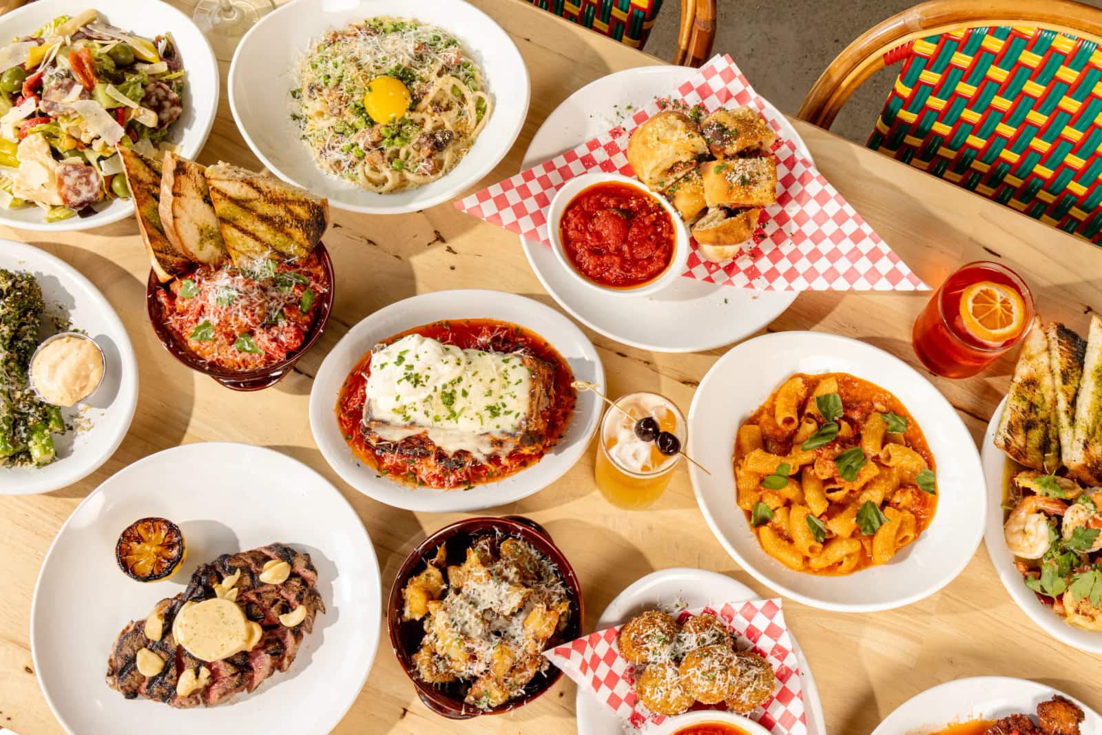 An overhead spread of Rosie's Italian dishes on a table — chicken parmesan, bucatini carbonara, rigatoni vodka, steak, stromboli, arancini and cocktails